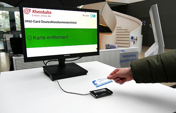 Eine Hand nimmt die HHU Card von einem Lesegerät, das vor einem Monitor steht
